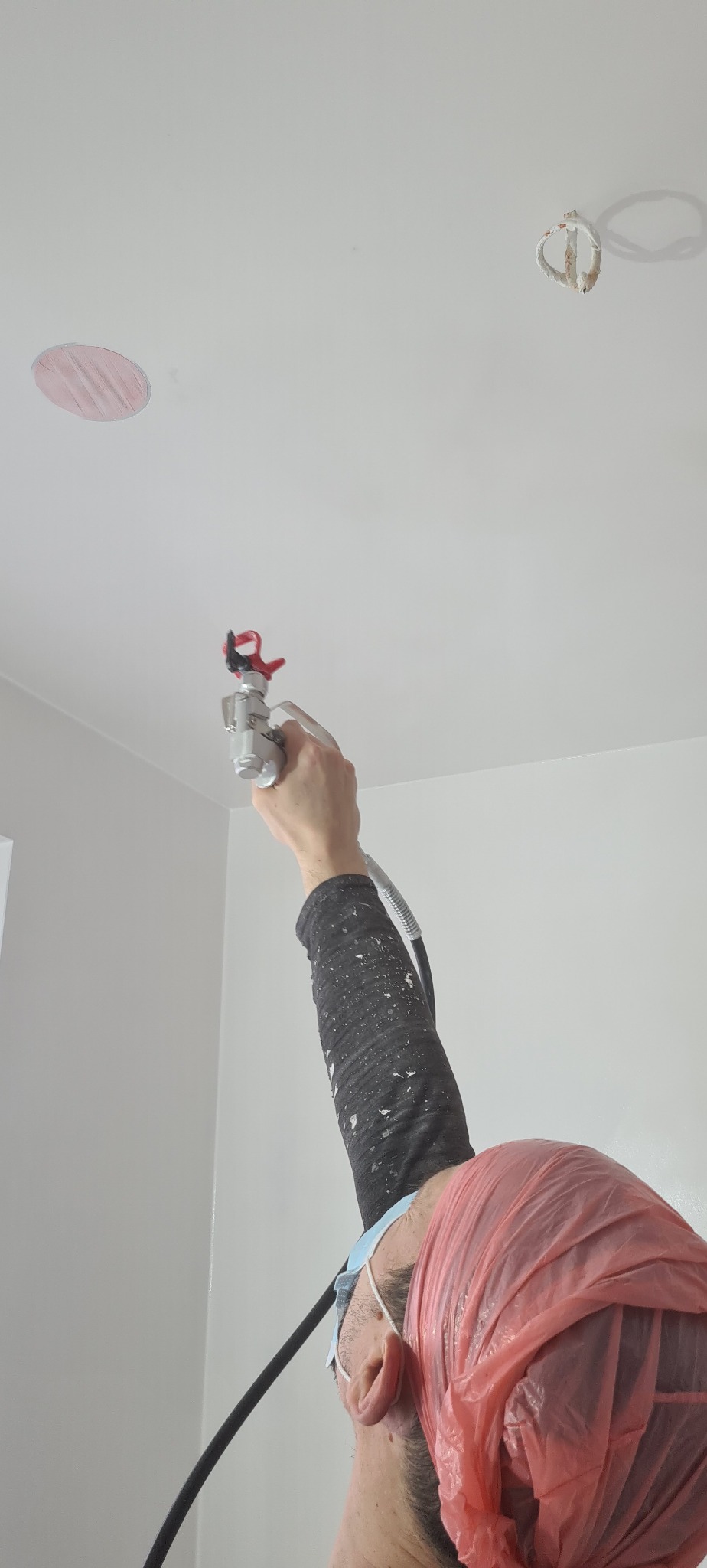 Malowanie natryskowe sufitu w pomieszczeniu, widoczny fragment twarzy malarza z zabezpieczeniem na głowie, pistolet natryskowy w dłoni oraz zaklejone miejsce po lampie.