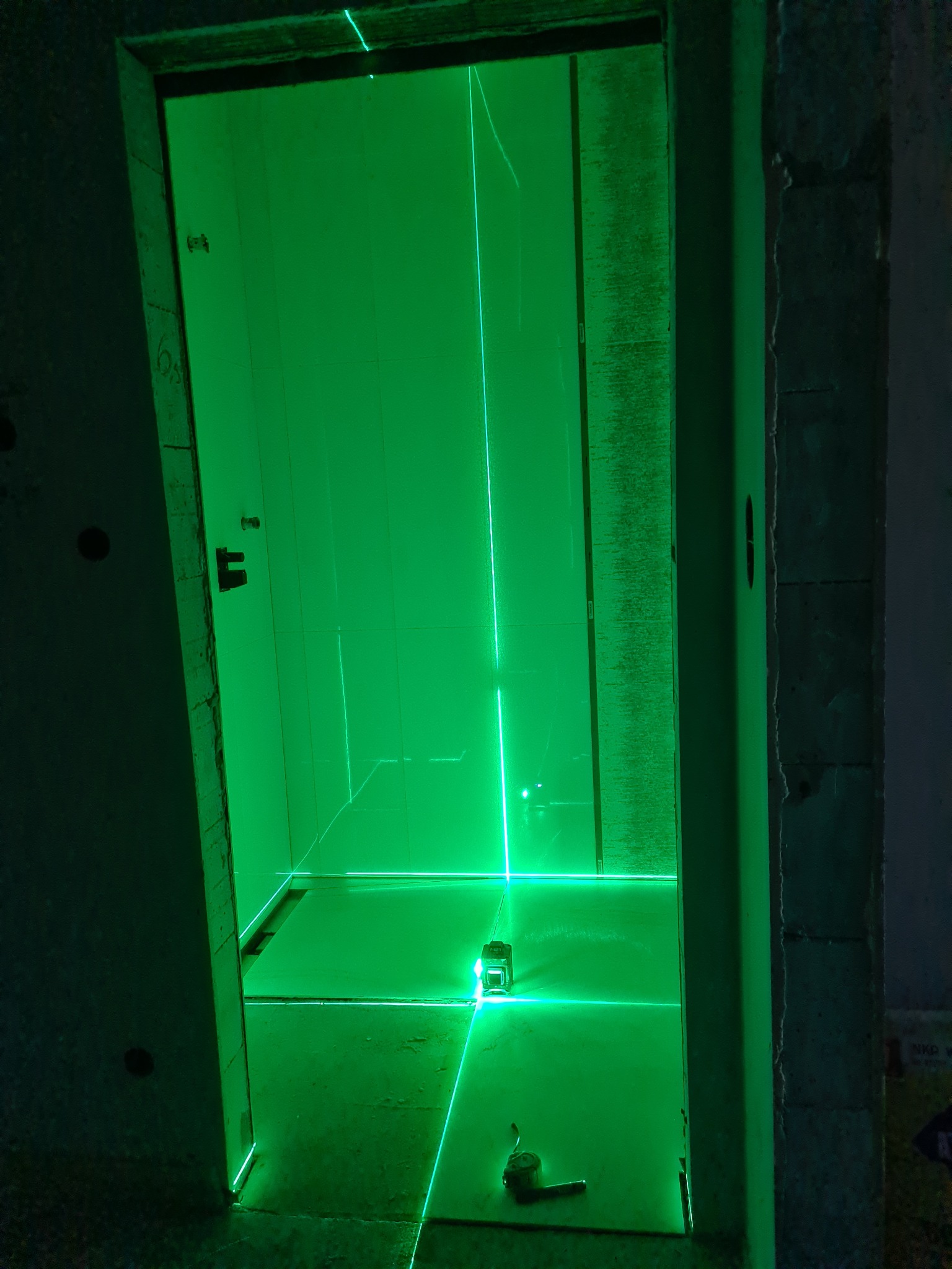 Wnętrze łazienki w trakcie remontu, oświetlone zielonym laserem krzyżowym, wyznaczającym poziomy na ścianach pokrytych płytkami i podłodze. Widoczny miernik laserowy i zwinięta miarka.