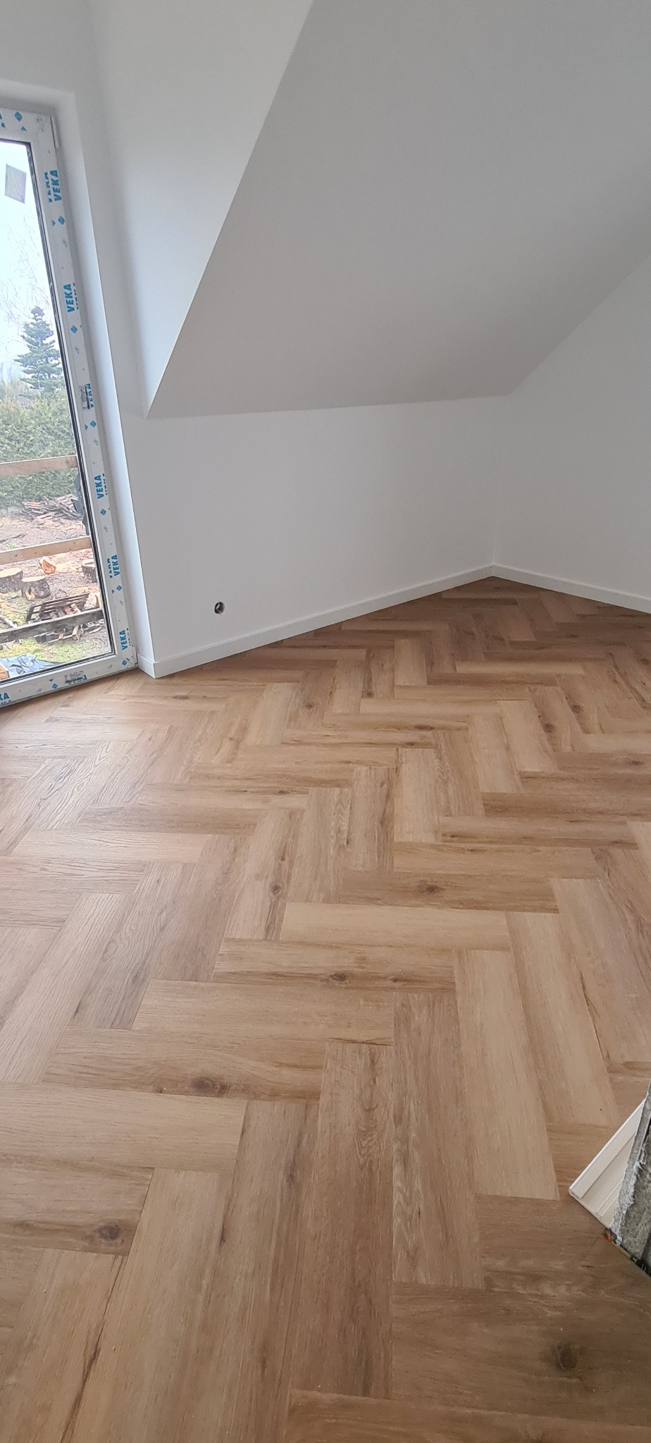 Jasne panele ułożone w jodełkę w pokoju na poddaszu z oknem i białymi ścianami.