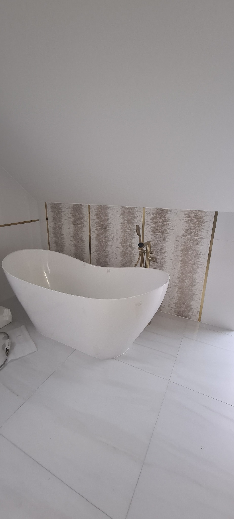 Wolnostojąca wanna w kształcie łodzi, umieszczona w łazience na poddaszu z białymi płytkami imitującymi marmur oraz dekoracyjnym panelem ściennym z poziomymi paskami w odcieniach beżu i złota...