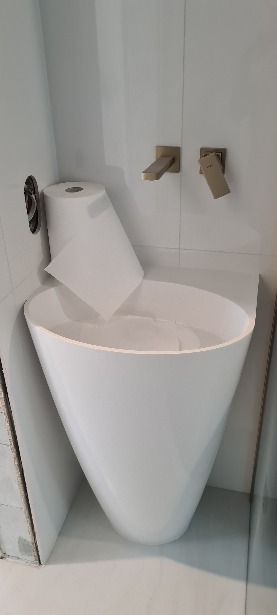 Nowoczesna, biała umywalka w kształcie stożka z nietypową, minimalistyczną baterią ścienną w kolorze szczotkowanego złota. Widoczny otwór w ścianie z wystającymi kablami elektrycznymi oraz rolka...