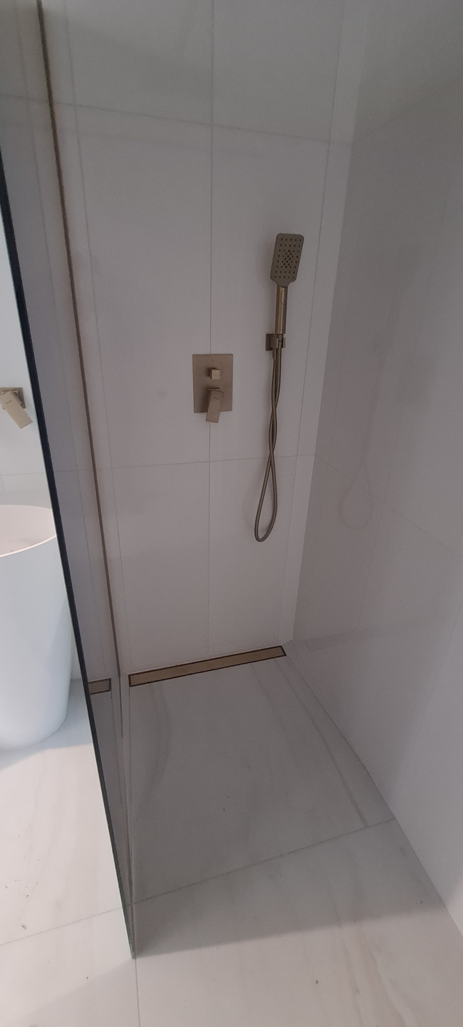 Nowoczesna kabina prysznicowa z odpływem liniowym i armaturą w odcieniach złota, białe płytki na ścianach, minimalistyczny design.