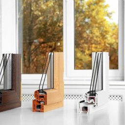 VIDOKOVO Robert Kania - Przekroje poprzeczne okien z trzema różnymi wykończeniami ramy (imitacja drewna ciemnego, jasnego i biała) prezentujące budowę wielokomorową i pakiet szybowy, na tle okna z widokiem na jesienne...