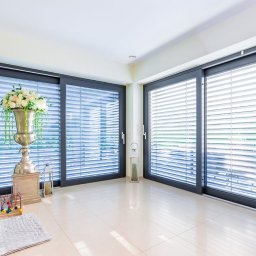 VIDOKOVO Robert Kania - Nowoczesne wnętrze z dużymi przesuwnymi oknami PCV w grafitowych ramach i wewnętrznymi poziomymi roletami, doświetlone naturalnym światłem, z dekoracyjną kompozycją kwiatową w srebrnej wazie...