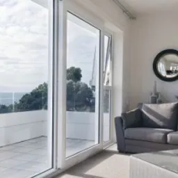 Jasne wnętrze salonu z dużym przesuwnym oknem balkonowym z białymi ramami, widok na taras z białymi balustradami i zieleń za nim, w tle morze, szara sofa i okrągłe lustro na ścianie.