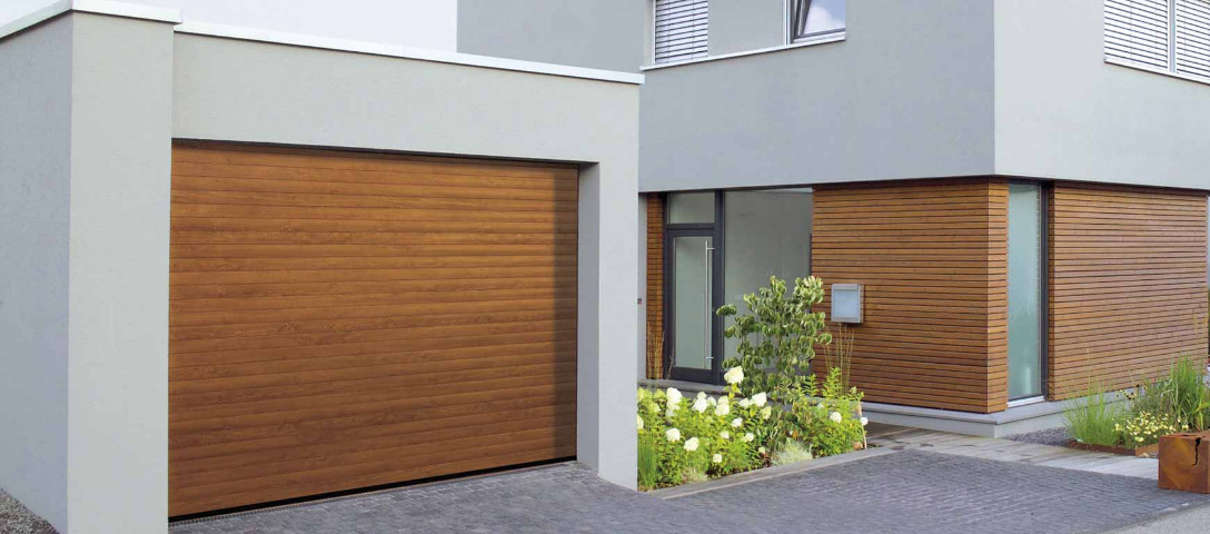 Nowoczesny dom z garażem i drewnianą bramą garażową segmentową w kolorze jasnego drewna, otoczony zadbaną zielenią.