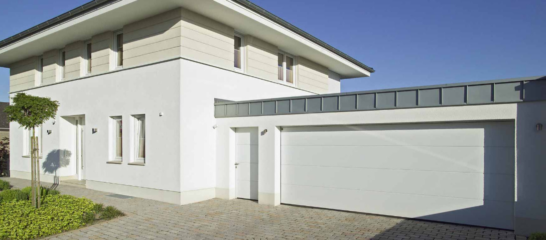 Nowoczesny dom z jasną elewacją i białą segmentową bramą garażową, widok z zewnątrz w słoneczny dzień.