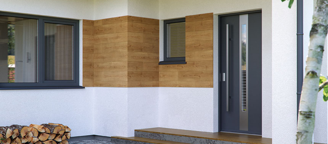 Elewacja domu z szarymi oknami, drzwiami wejściowymi z przeszkleniami i okładziną imitującą drewno, stos drewna opałowego pod oknem.
