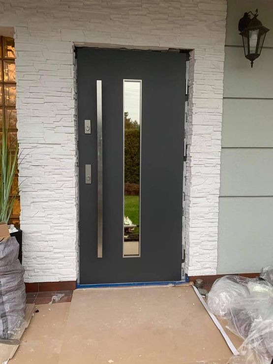 Grafitowe drzwi wejściowe z pionowym przeszkleniem i długim, stalowym uchwytem, zamontowane w elewacji z białej, dekoracyjnej cegły, widoczne ślady prac montażowych.