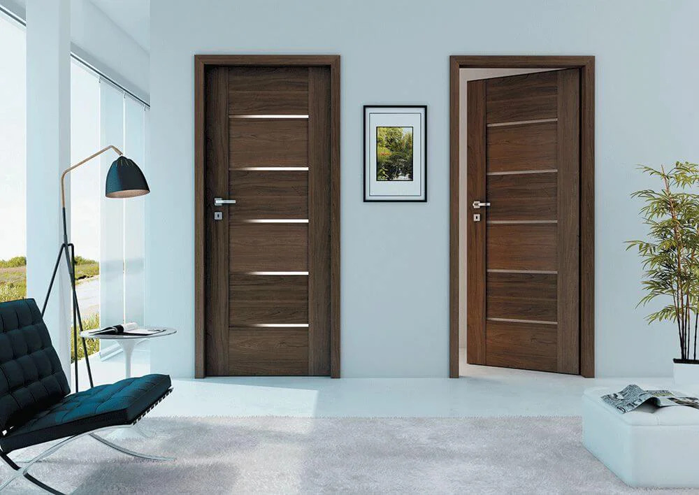 Dwie nowoczesne, brązowe drzwi z poziomymi, jasnymi wstawkami w minimalistycznym wnętrzu z czarnym fotelem, lampą i rośliną doniczkową.