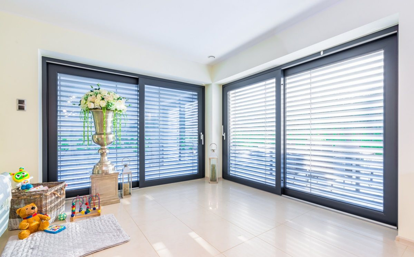 Nowoczesne wnętrze z dużymi przesuwnymi oknami PCV w grafitowych ramach i wewnętrznymi poziomymi roletami, doświetlone naturalnym światłem, z dekoracyjną kompozycją kwiatową w srebrnej wazie...