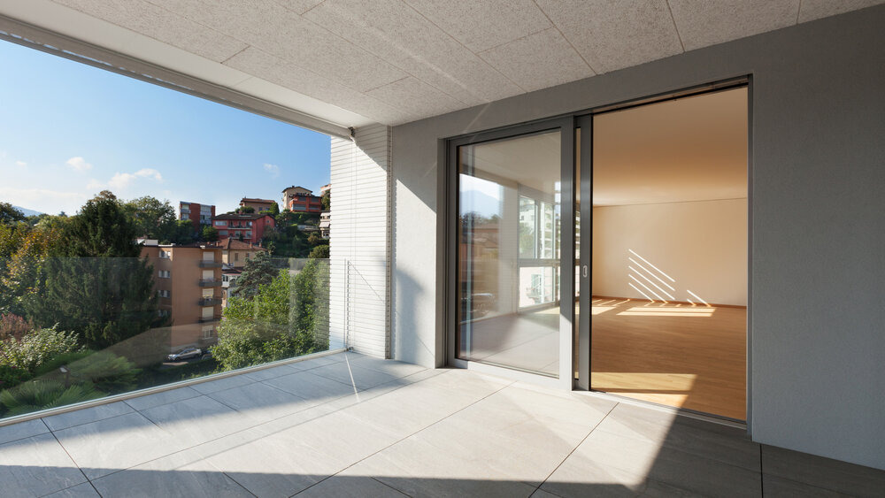 Nowoczesny balkon z szarymi płytkami podłogowymi i przesuwnymi drzwiami balkonowymi z widokiem na zieleń i zabudowę miejską.