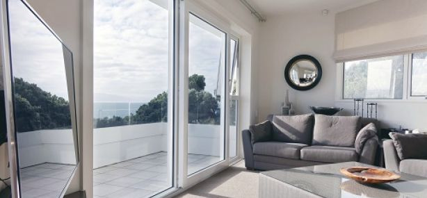 Jasne wnętrze salonu z dużym przesuwnym oknem balkonowym z białymi ramami, widok na taras z białymi balustradami i zieleń za nim, w tle morze, szara sofa i okrągłe lustro na ścianie.
