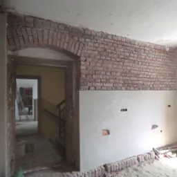 Wnętrze w trakcie remontu: fragment ściany z ceglanym łukiem nad przejściem, dolna część ściany pokryta świeżym tynkiem, podłoga zasypana piaskiem.