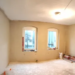 Wnętrze pokoju po nałożeniu świeżych, jasnych tynków na ścianach i suficie, z widocznymi dwoma oknami o zaokrąglonych górnych krawędziach, pozostawionymi narzędziami i materiałami budowlanymi...