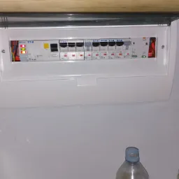 Otwarta skrzynka bezpiecznikowa marki Eaton z widocznymi bezpiecznikami i przewodami. Skrzynka zamontowana na ścianie, pod drewnianą półką. Na dole butelka.