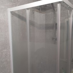 SZYMO TRANS SPÓŁKA Z OGRANICZONĄ ODPOWIEDZIALNOŚCIĄ - Szklana kabina prysznicowa z deszczownicą, wykończona w szarych tonacjach. Widoczne detale armatury i płytek. Minimalistyczny design.