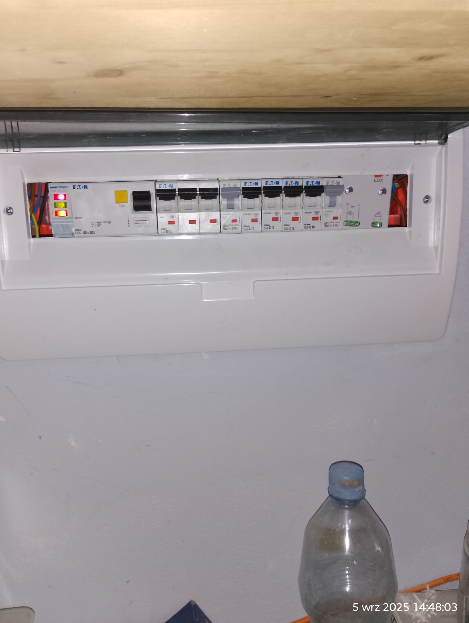 Otwarta skrzynka bezpiecznikowa marki Eaton z widocznymi bezpiecznikami i przewodami. Skrzynka zamontowana na ścianie, pod drewnianą półką. Na dole butelka.