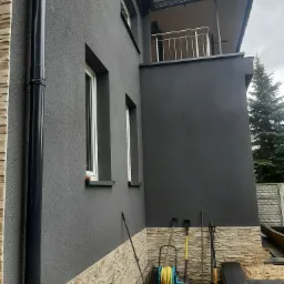 Szara elewacja domu z elementami dekoracyjnymi z kamienia, widoczna rynna, okna i narzędzia ogrodnicze przy ścianie.