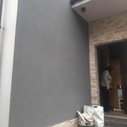 Tom-Bud - Szara elewacja budynku z dekoracyjnym wykończeniem z kamienia, widoczne worki i wiadro z resztkami materiałów budowlanych, kobieta wchodząca do budynku.