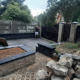 Tom-Bud - Układanie kostki brukowej na podwórku z widocznymi elementami muru i ogrodzenia, w tle osoba robiąca zdjęcie telefonem.