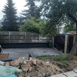 Tom-Bud - Układanie kostki brukowej na podwórku, widoczny fragment nowej nawierzchni, przygotowane podłoże pod rabatę i stos kamieni oraz ziemi, w tle ogrodzenie z betonowych elementów.