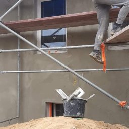 Tom-Bud - Pracownik na rusztowaniu podczas nakładania szarego tynku na elewację budynku, widoczny kubeł z zaprawą na stercie piasku, okna zabezpieczone folią.