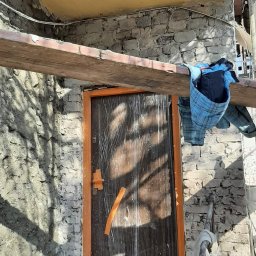 Tom-Bud - Wejście do budynku w trakcie prac remontowych, zabezpieczone folią drzwi z pomarańczową futryną, widoczne rusztowanie, fragment ocieplonego sufitu i ubranie robocze na belce.