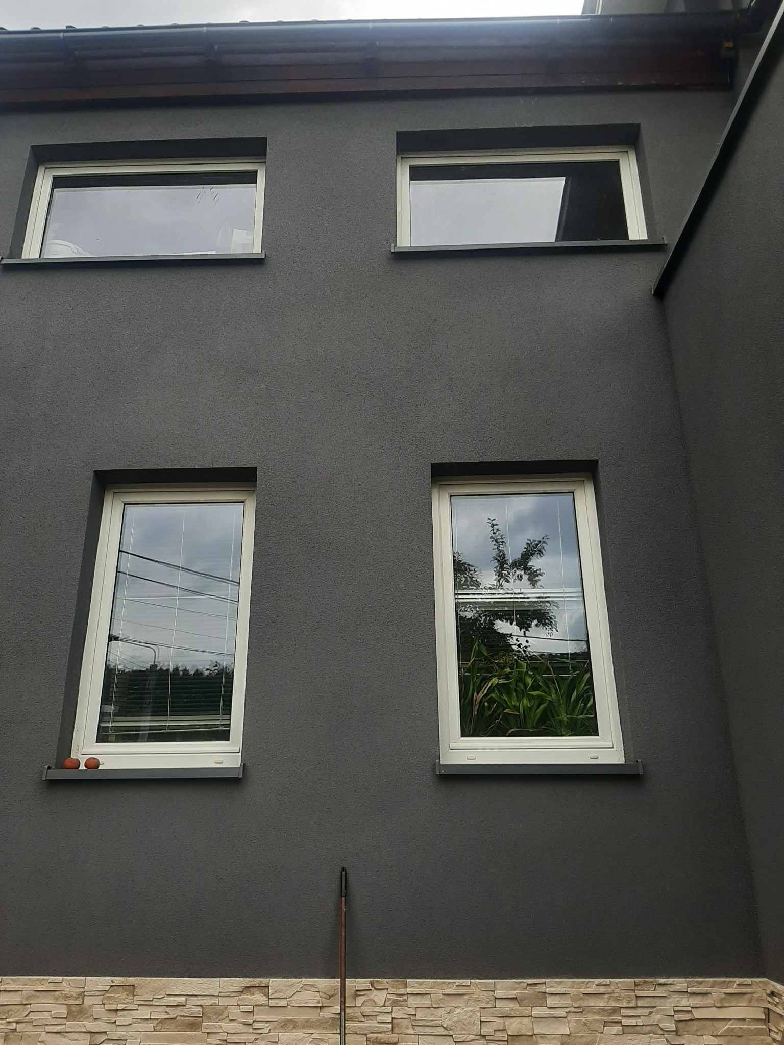 Szara elewacja budynku z czterema oknami w białych ramach, dolna część wykończona kamieniem dekoracyjnym, na parapecie dwa pomarańczowe obiekty.