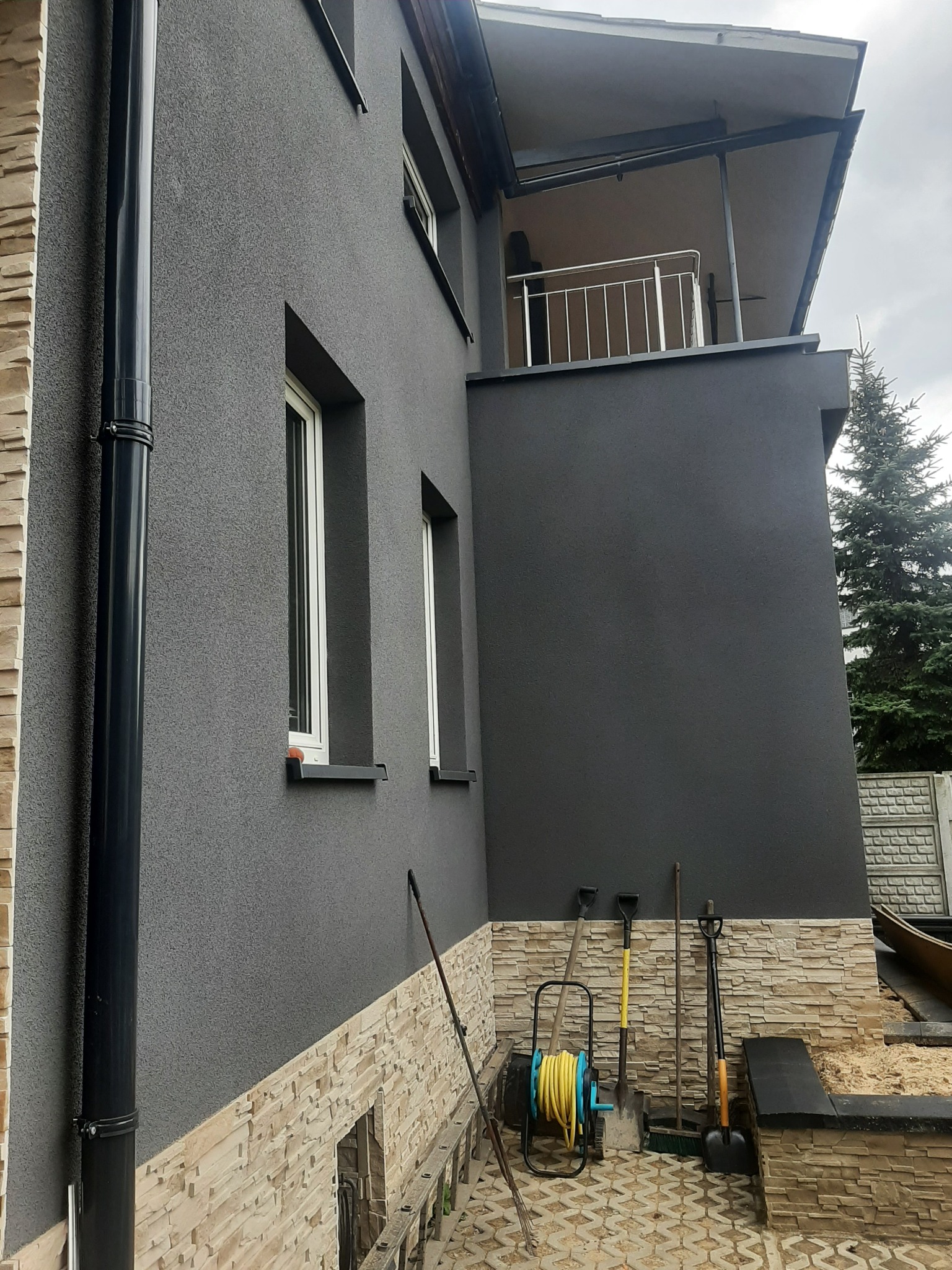 Szara elewacja domu z elementami dekoracyjnymi z kamienia, widoczna rynna, okna i narzędzia ogrodnicze przy ścianie.