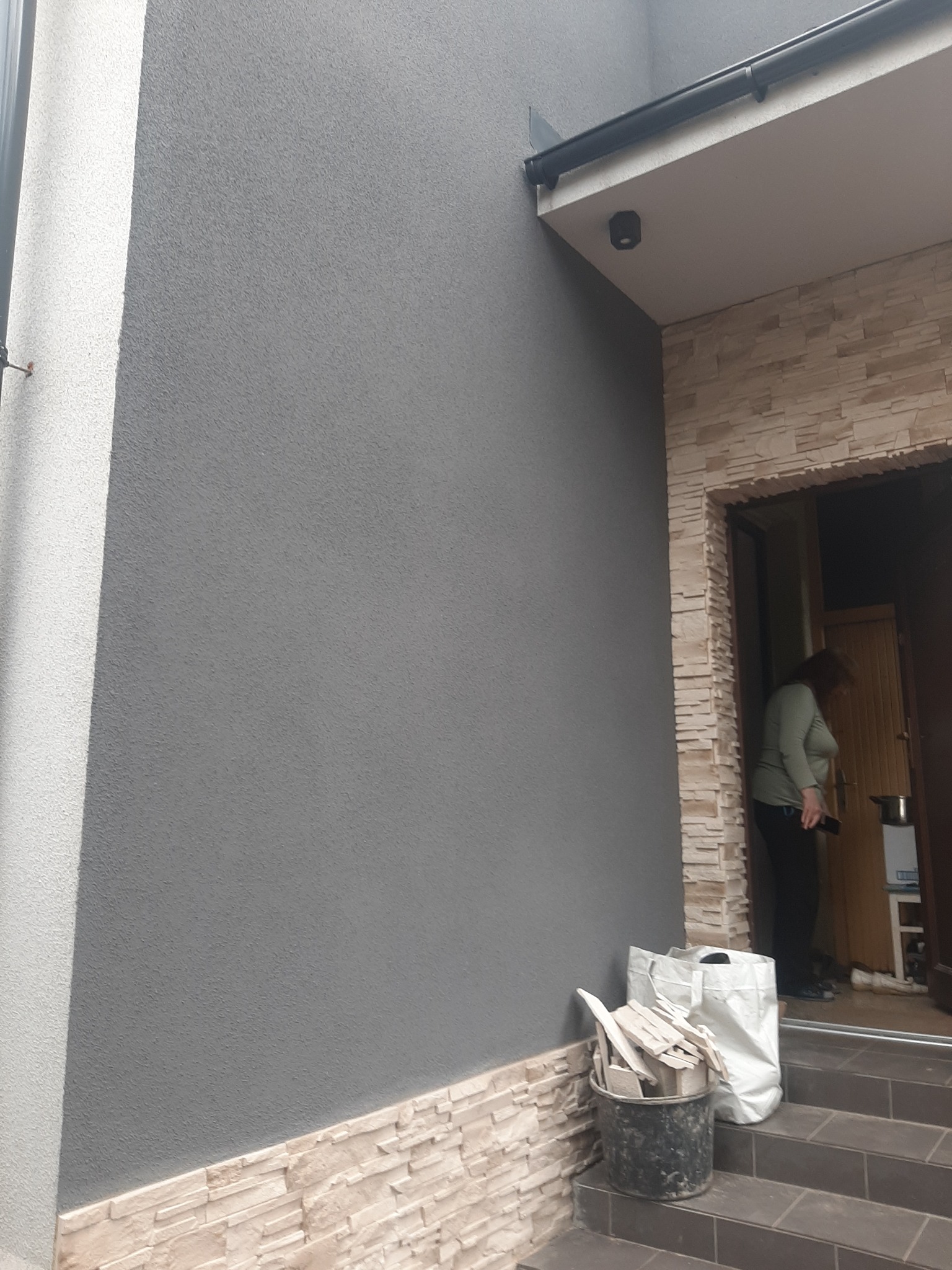 Szara elewacja budynku z dekoracyjnym wykończeniem z kamienia, widoczne worki i wiadro z resztkami materiałów budowlanych, kobieta wchodząca do budynku.
