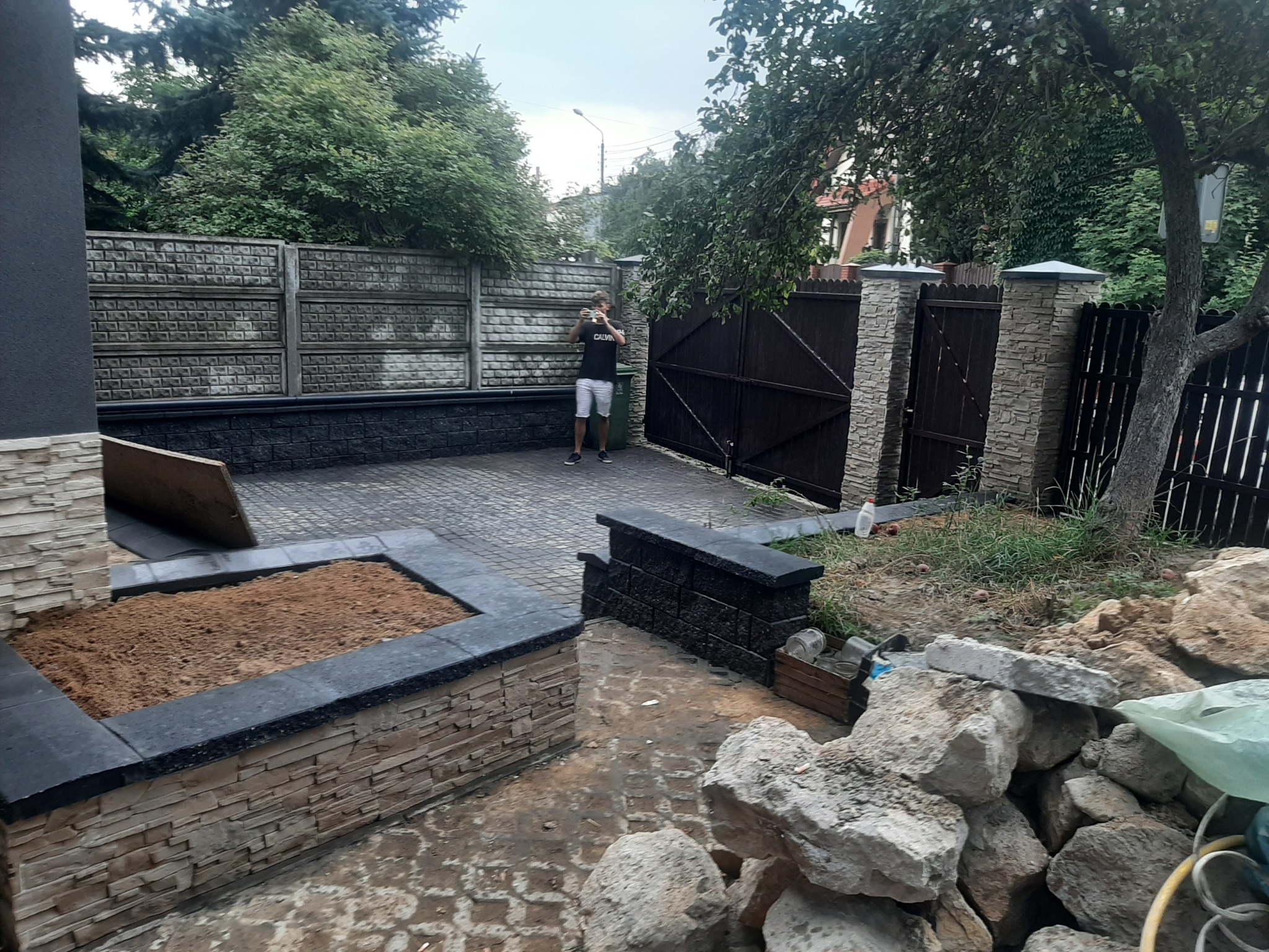 Układanie kostki brukowej na podwórku z widocznymi elementami muru i ogrodzenia, w tle osoba robiąca zdjęcie telefonem.