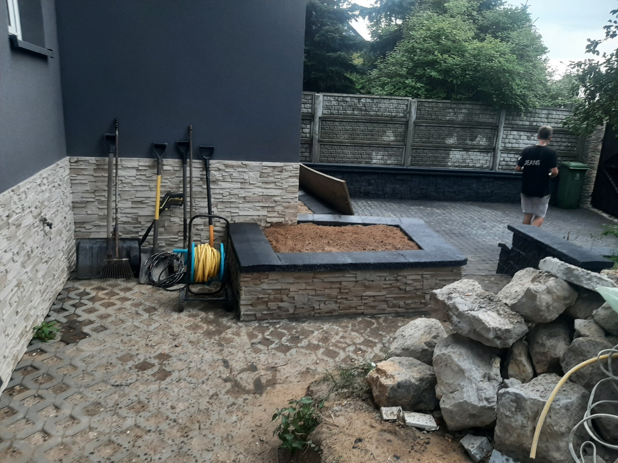 Budowa podwyższonej rabaty ogrodowej z kamienia, z narzędziami ogrodniczymi opartymi o ścianę budynku, widok na betonowy płot i drzewa w tle
