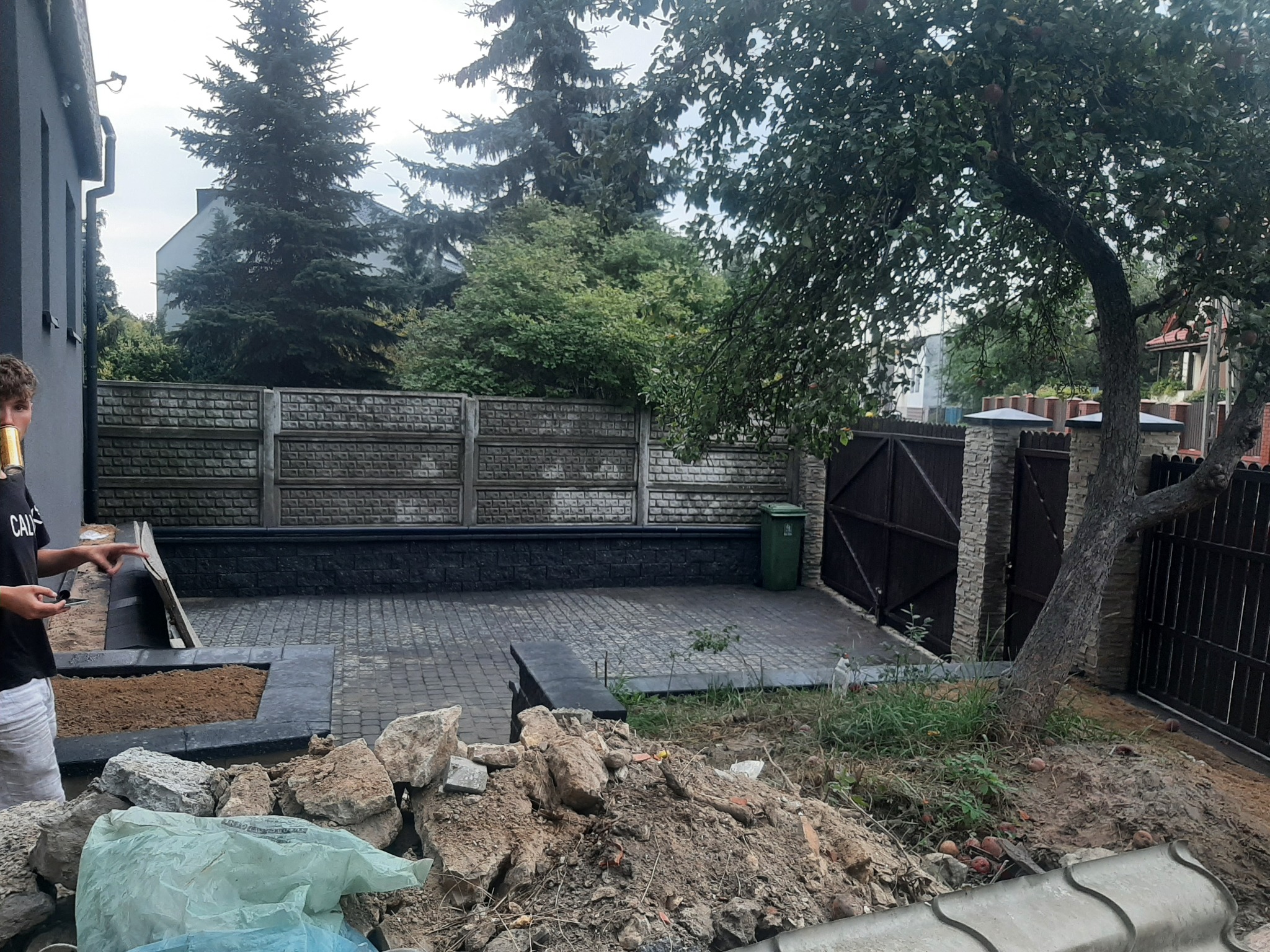 Układanie kostki brukowej na podwórku, widoczny fragment nowej nawierzchni, przygotowane podłoże pod rabatę i stos kamieni oraz ziemi, w tle ogrodzenie z betonowych elementów.