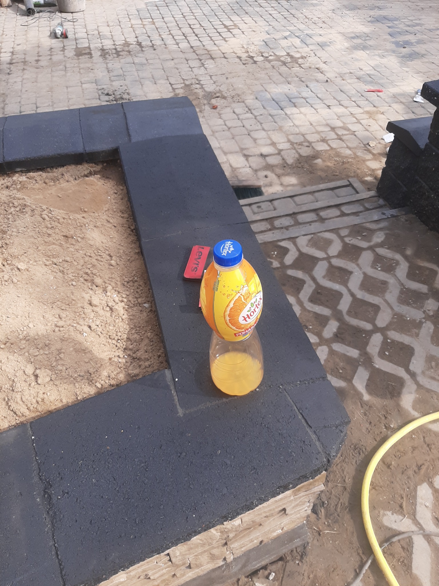 Krawężnik z ciemnych kostek obrzeżający klomb z piaskiem; butelka soku Hortex i czerwona packa do fugowania na krawężniku. W tle kostka brukowa i zwój żółtego węża.