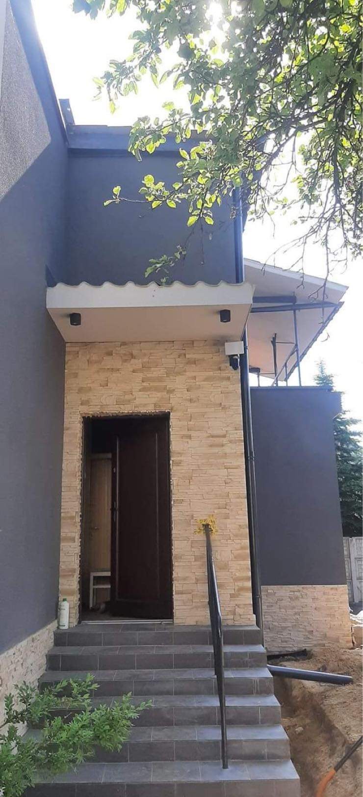 Wejście do domu z elewacją z kamienia dekoracyjnego, schody wyłożone szarą płytką, widoczny fragment dachu i rynny, czarna poręcz i pędzel do malowania oparty o ścianę.