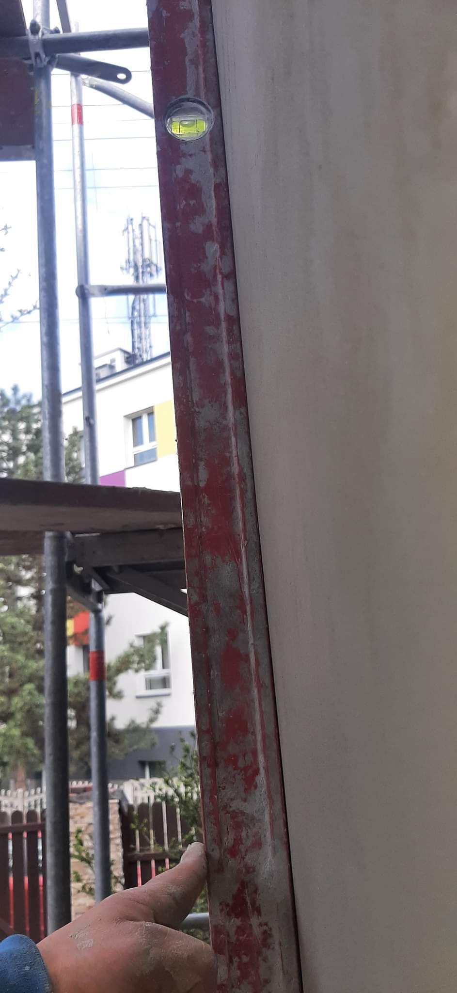 Sprawdzanie pionu ściany za pomocą poziomicy na rusztowaniu, w tle budynek mieszkalny.