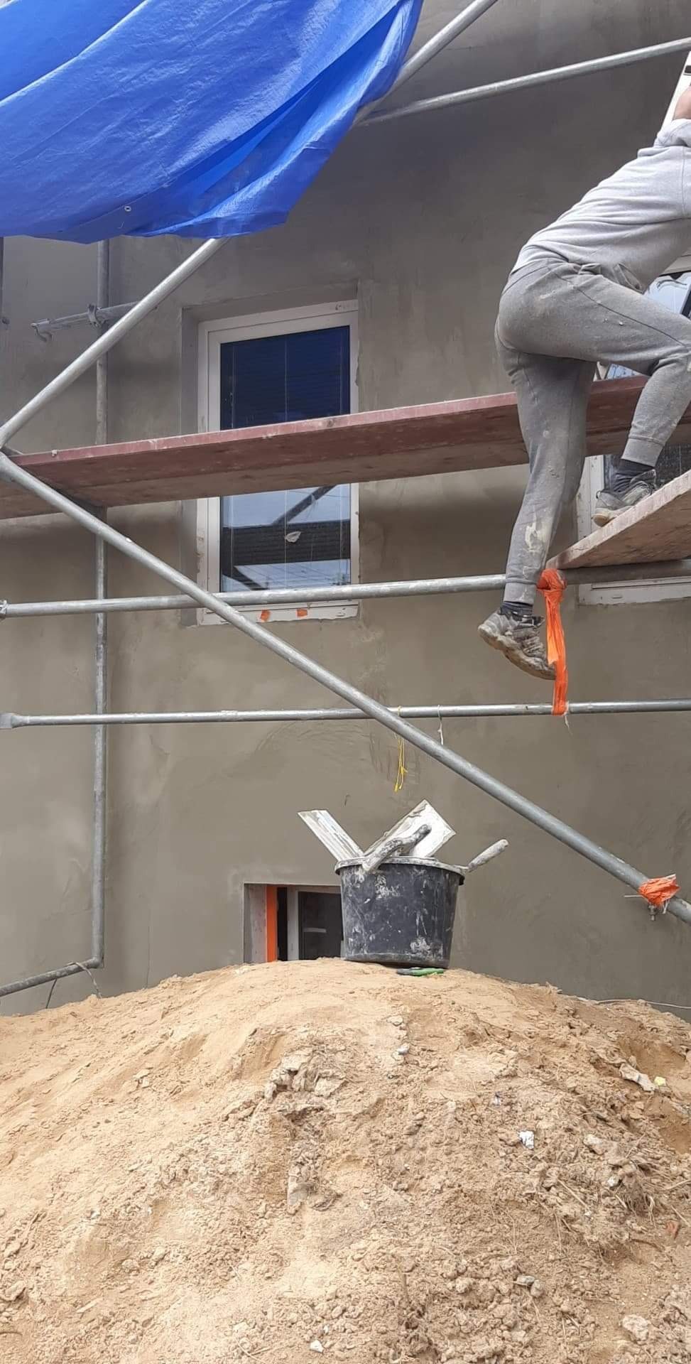 Pracownik na rusztowaniu podczas nakładania szarego tynku na elewację budynku, widoczny kubeł z zaprawą na stercie piasku, okna zabezpieczone folią.