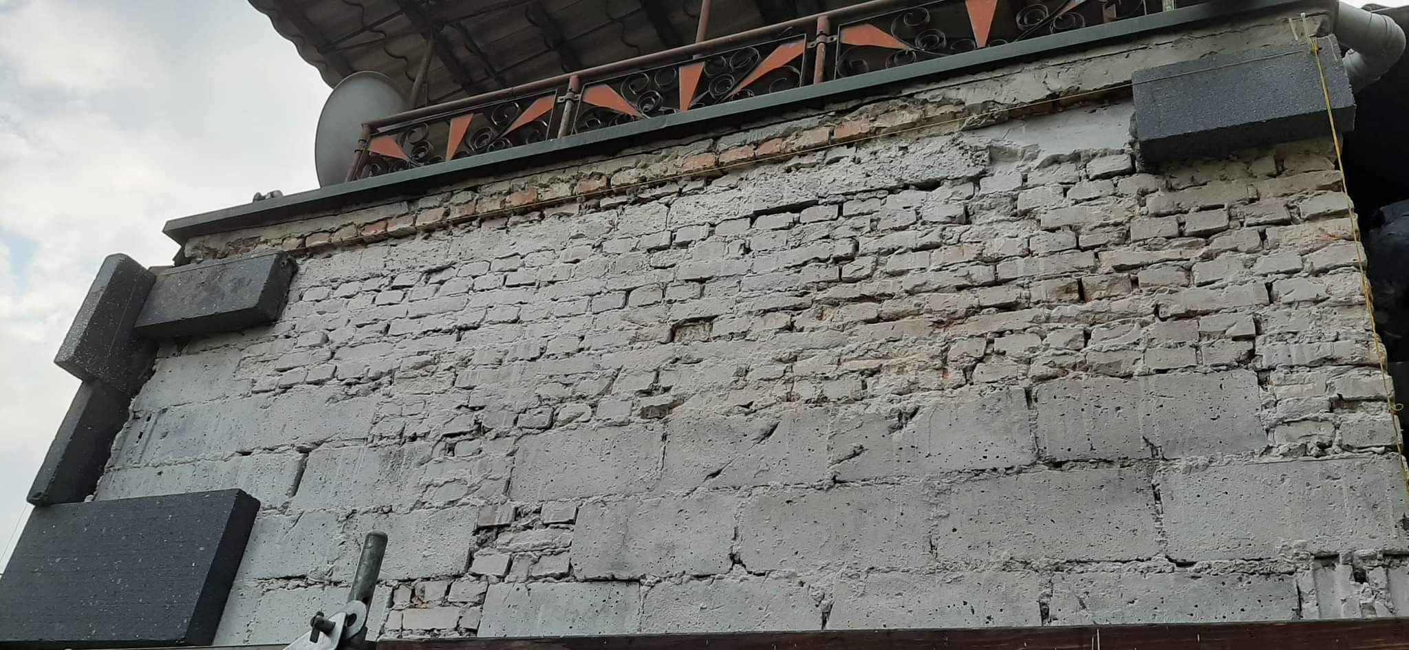 Fragment ściany z cegły i betonu z widocznymi fragmentami ocieplenia z szarego styropianu, z góry widoczna balustrada z metaloplastyki.