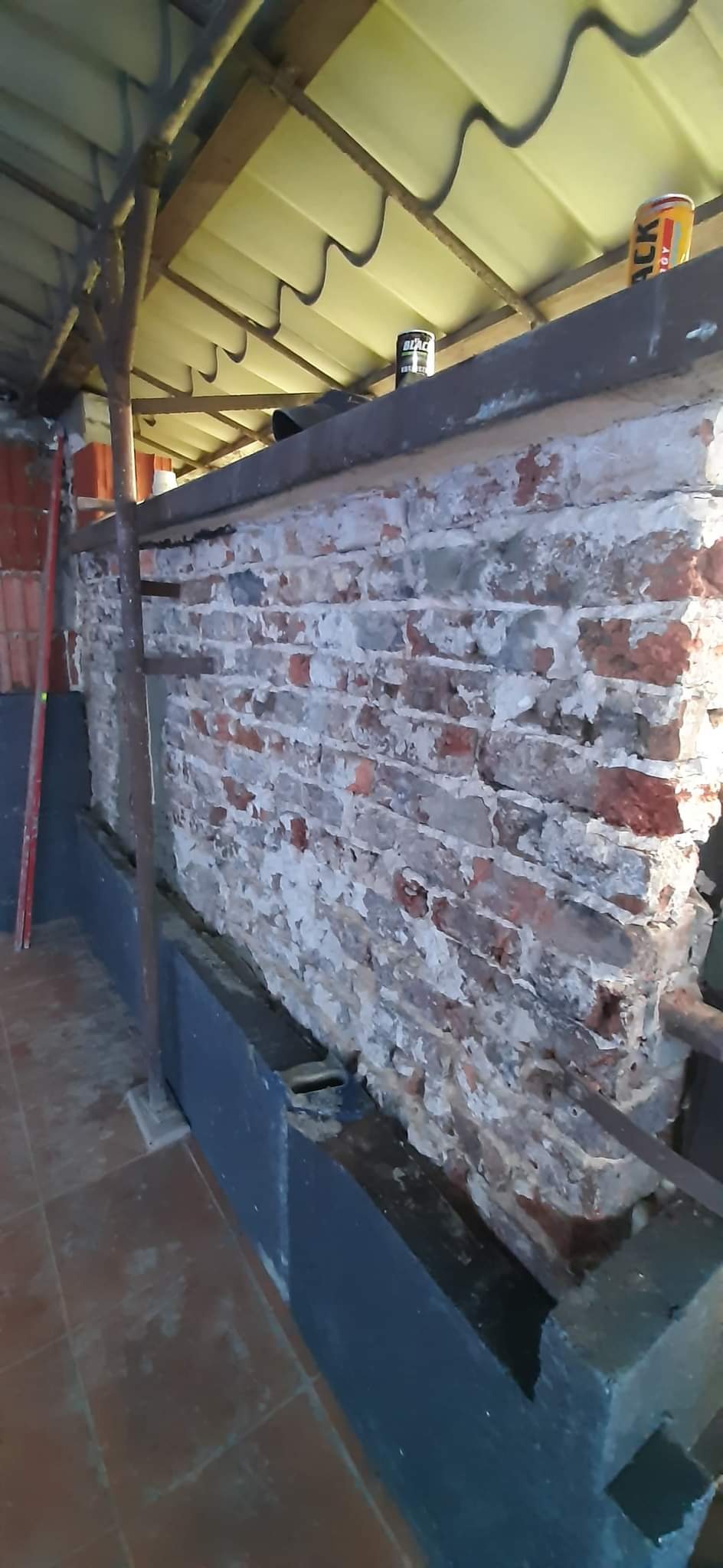 Fragment ściany z czerwonej cegły z resztkami białego tynku, wspartej na stalowych profilach, pod zadaszeniem z blachy trapezowej, widoczne puszki z napojami na górze ściany.