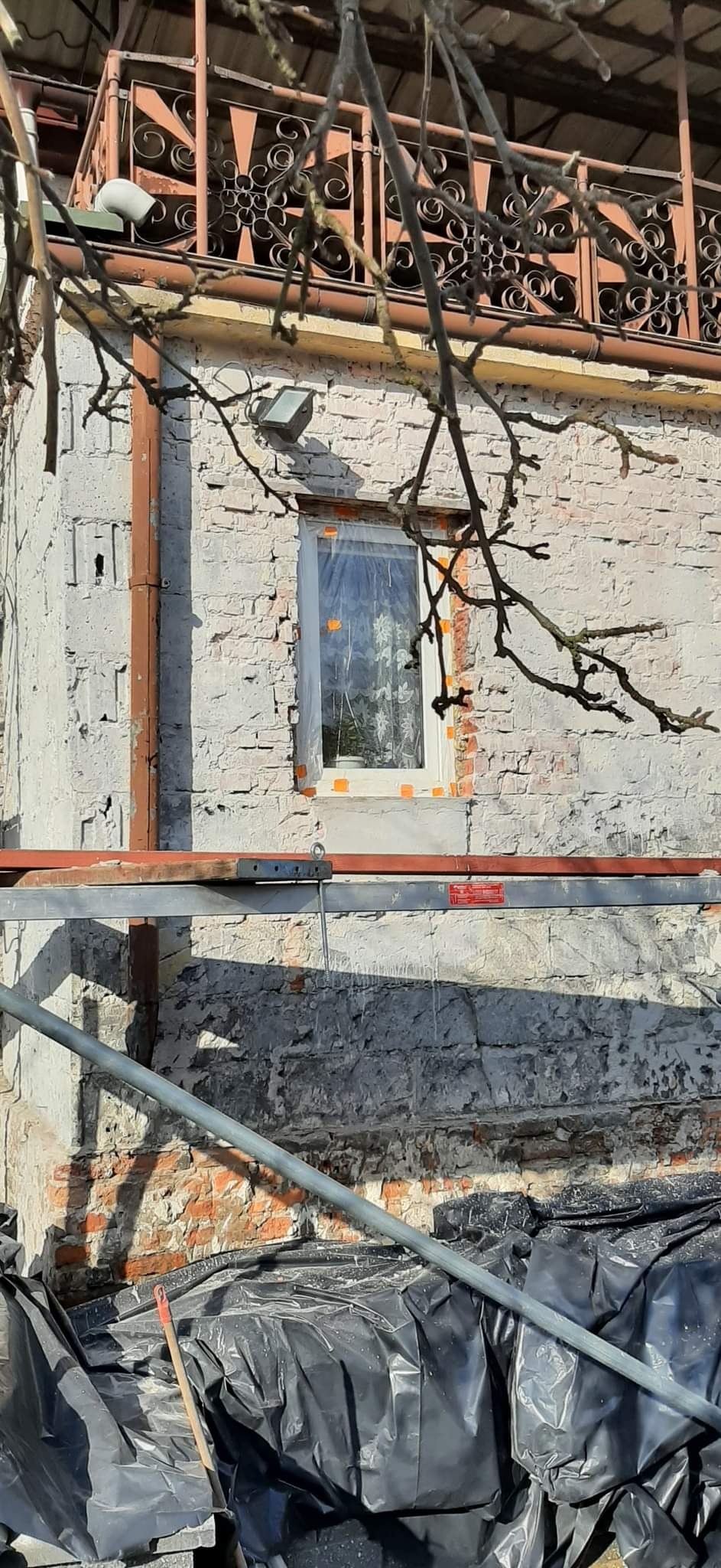 Fragment elewacji budynku z oknem zabezpieczonym folią malarską, widoczne rusztowanie i gałęzie drzewa na pierwszym planie, ślady prac murarskich na ścianie.
