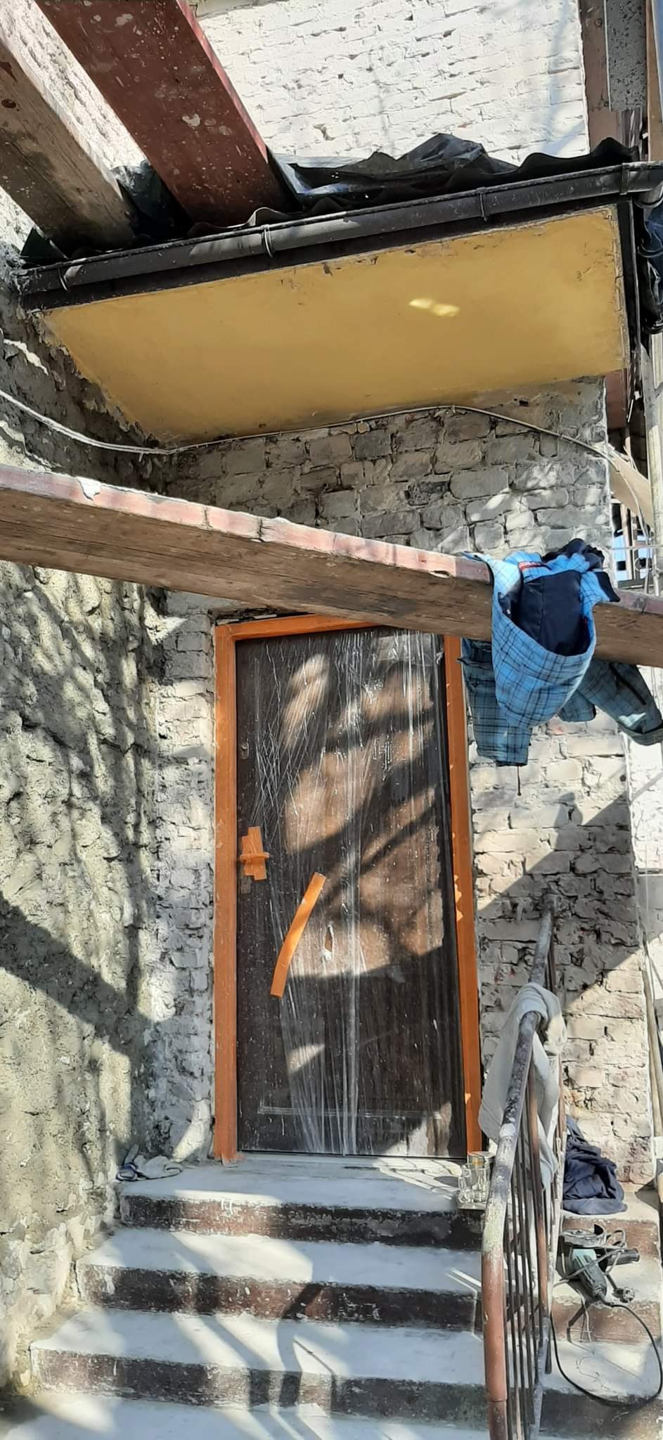 Wejście do budynku w trakcie prac remontowych, zabezpieczone folią drzwi z pomarańczową futryną, widoczne rusztowanie, fragment ocieplonego sufitu i ubranie robocze na belce.