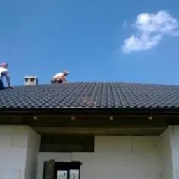 Montaż czarnej dachówki na nowym domu z białego pustaka. Dwóch dekarzy w kaskach pracuje na dachu w słoneczny dzień. Widok z dołu.