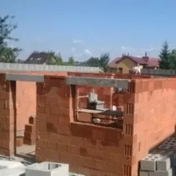 Budowa domu jednorodzinnego w trakcie: ściany z czerwonej cegły, nadproża betonowe, widoczne okna i drzwi. Na murze stoi osoba. W tle domy i zieleń.