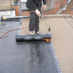 Janusz Kowalski - Dekarz w trakcie pracy z papą termozgrzewalną na płaskim dachu; palnik w dłoni, rolka papy z nadrukiem logo, widoczne sąsiednie budynki.