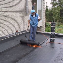 Janusz Kowalski - Dekarz z palnikiem podczas hydroizolacji dachu papą termozgrzewalną, widoczny mur z cegły i osoba w oknie.