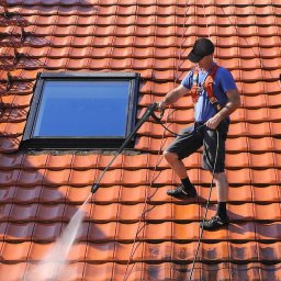 Janusz Kowalski - Pracownik w uprzęży, z czapką na głowie, czyści czerwoną dachówkę z okienkiem dachowym za pomocą myjki ciśnieniowej, strumień wody usuwa zabrudzenia.