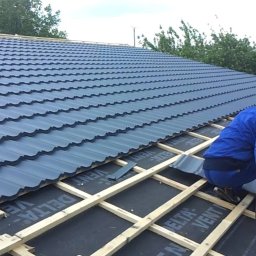 Janusz Kowalski - Monter w niebieskim kombinezonie przycina blachodachówkę szlifierką kątową na drewnianej konstrukcji dachu pokrytej czarną membraną, w tle widoczny fragment gotowego pokrycia.