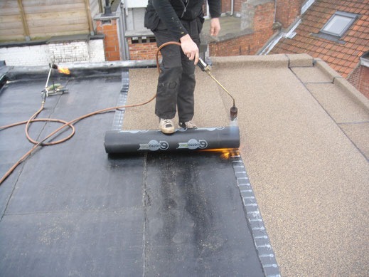 Dekarz w trakcie pracy z papą termozgrzewalną na płaskim dachu; palnik w dłoni, rolka papy z nadrukiem logo, widoczne sąsiednie budynki.