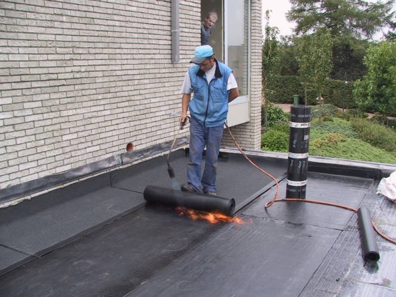 Dekarz z palnikiem podczas hydroizolacji dachu papą termozgrzewalną, widoczny mur z cegły i osoba w oknie.